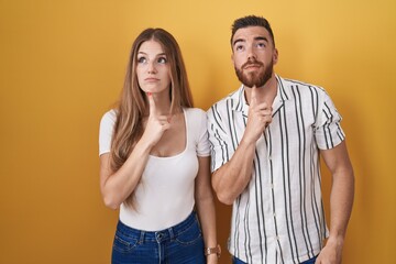 Young couple standing over yellow background thinking concentrated about doubt with finger on chin and looking up wondering