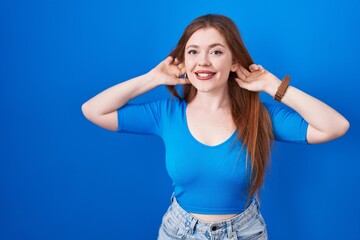 Obraz na płótnie Canvas Redhead woman standing over blue background smiling pulling ears with fingers, funny gesture. audition problem