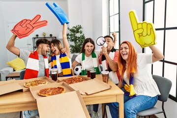 Group of young friends watching and supporting soccer match eating pizza at home.