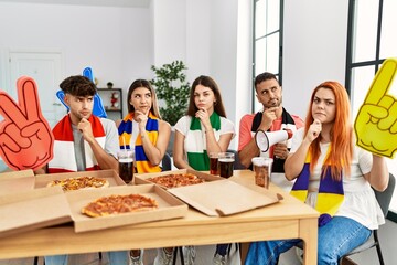 Group of young hispanic people eating pizza supporting soccer team at home serious face thinking about question with hand on chin, thoughtful about confusing idea