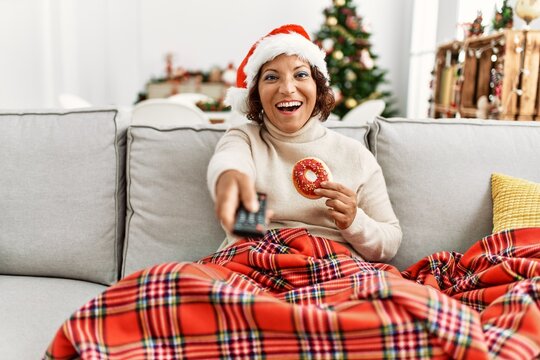 Middle age hispanic woman wearing christmas hat watching movie sitting on the sofa at home.