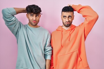 Young hispanic gay couple standing over pink background confuse and wondering about question....