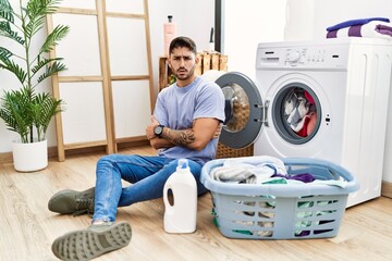Young hispanic man putting dirty laundry into washing machine skeptic and nervous, disapproving expression on face with crossed arms. negative person.