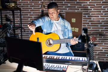 Hispanic young man playing classic guitar at music studio looking unhappy and angry showing rejection and negative with thumbs down gesture. bad expression.