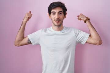 Young hispanic man standing over pink background showing arms muscles smiling proud. fitness concept.