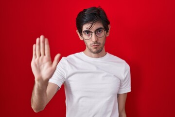 Young hispanic man standing over red background doing stop sing with palm of the hand. warning...