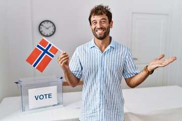 Young handsome man at political campaign election holding norway flag celebrating achievement with happy smile and winner expression with raised hand