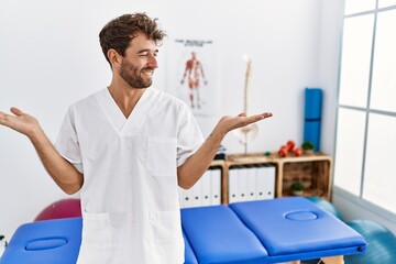 Young handsome physiotherapist man working at pain recovery clinic smiling showing both hands open palms, presenting and advertising comparison and balance