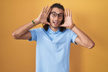 Fototapeta na wymiar Young hispanic man standing over yellow background smiling cheerful playing peek a boo with hands showing face. surprised and exited