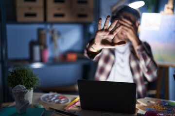 Young hispanic man sitting at art studio with laptop late at night covering eyes with hands and doing stop gesture with sad and fear expression. embarrassed and negative concept.