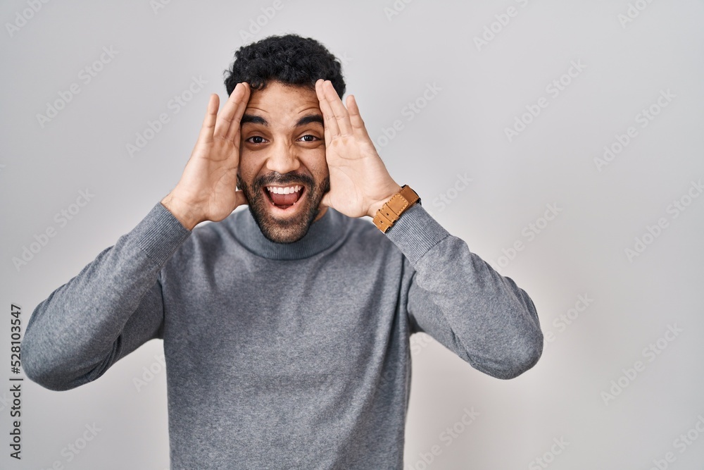 Sticker Hispanic man with beard standing over white background smiling cheerful playing peek a boo with hands showing face. surprised and exited