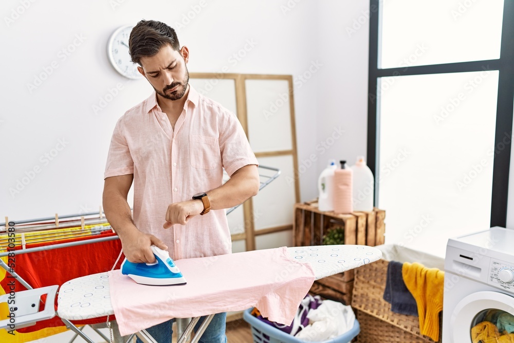 Poster Young man with beard ironing clothes at home checking the time on wrist watch, relaxed and confident