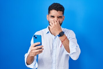 Hispanic man with beard using smartphone typing message smelling something stinky and disgusting, intolerable smell, holding breath with fingers on nose. bad smell