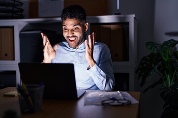 Hispanic man with beard working at the office with laptop at night celebrating victory with happy smile and winner expression with raised hands