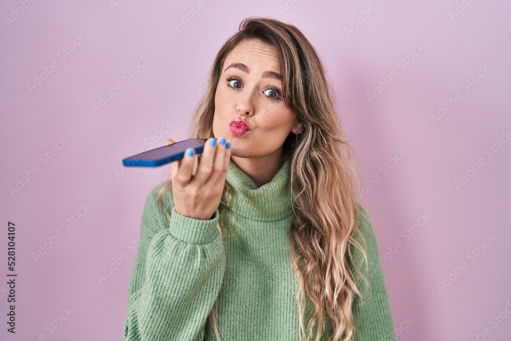 Poster Young caucasian woman sending a voice message with smartphone looking at the camera blowing a kiss being lovely and sexy. love expression.