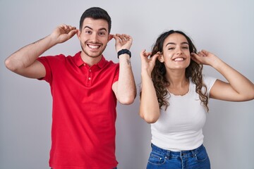 Young hispanic couple standing over isolated background smiling pulling ears with fingers, funny...