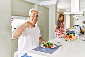 Middle age hispanic couple eating healthy meal at home pointing finger to one self smiling happy and proud