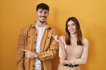 Young hispanic couple standing over yellow background with a big smile on face, pointing with hand finger to the side looking at the camera.