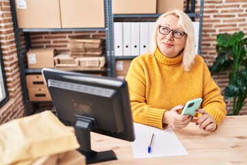 Middle age blonde woman ecommerce business worker using smartphone at office