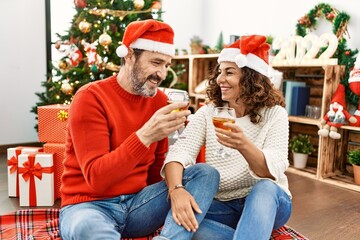 Middle age hispanic couple wearing christmas hat. Sitting on the floor toasting with champagne at home.