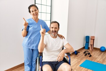 Obraz premium Hispanic middle age man sitting on wheelchair and nurse at rehabilitation clinic smiling happy pointing with hand and finger to the side