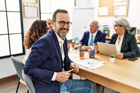 Group of middle age business workers working at the office. Man smiling happy and looking to the camera sitting on the table.
