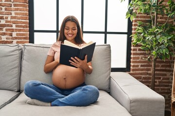 Young latin woman pregnant touching belly reading book at home