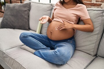 Young latin woman pregnant applying cream lotion on skin belly at home