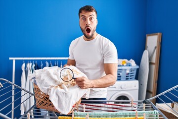Handsome hispanic man holding magnifying glass looking for stain at clothes afraid and shocked with surprise and amazed expression, fear and excited face.