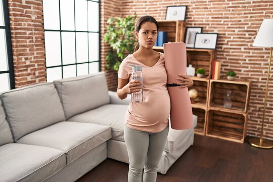 Young pregnant woman holding yoga mat at home looking at the camera blowing a kiss being lovely and sexy. love expression.