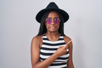 Young african american with braids wearing hat and sunglasses pointing aside worried and nervous with forefinger, concerned and surprised expression
