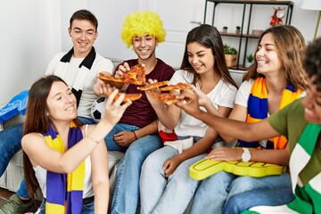 Group of young friends watching and supporting soccer match eating pizza at home.