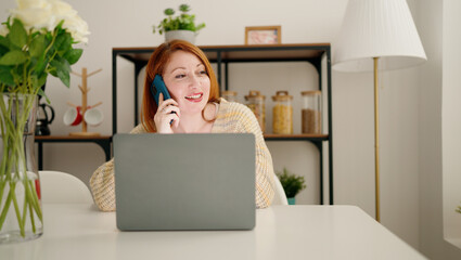 Young redhead woman using laptop and talking on the smartphone at home