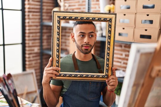 Young hispanic man sitting at art studio with empty frame smiling looking to the side and staring away thinking.