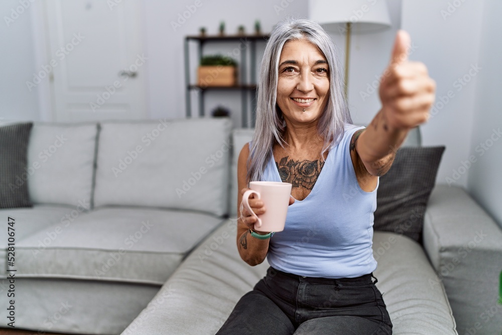 Wall mural middle age grey-haired woman drinking coffee sitting on the sofa at home approving doing positive ge