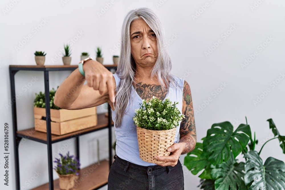 Sticker Middle age grey-haired woman holding green plant pot at home pointing down looking sad and upset, indicating direction with fingers, unhappy and depressed.