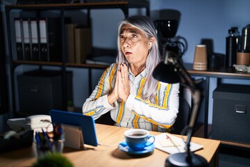 Middle age woman with grey hair working at the office at night begging and praying with hands together with hope expression on face very emotional and worried. begging.