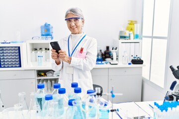 Middle age grey-haired woman wearing scientist uniform using smartphone at laboratory