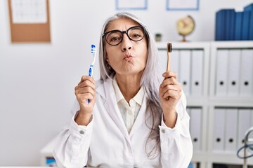 Middle age grey-haired woman working at dentist clinic holding electric and recycled teethbrush looking at the camera blowing a kiss being lovely and sexy. love expression.