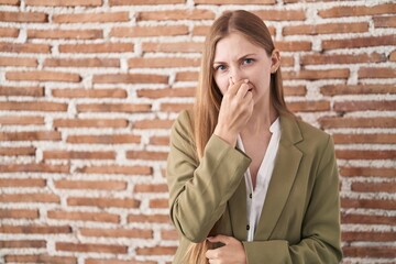 Young caucasian woman standing over bricks wall background smelling something stinky and disgusting, intolerable smell, holding breath with fingers on nose. bad smell