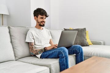 Young hispanic man using laptop sitting on the sofa at home.