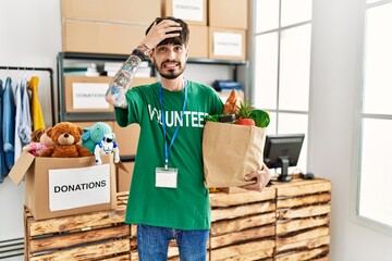 Hispanic man with beard wearing volunteer t shirt at donations point stressed and frustrated with hand on head, surprised and angry face