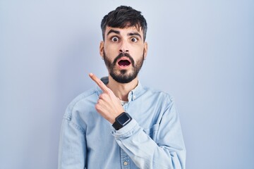 Young hispanic man with beard standing over blue background surprised pointing with finger to the side, open mouth amazed expression.