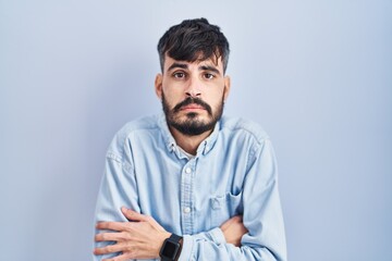 Young hispanic man with beard standing over blue background shaking and freezing for winter cold...