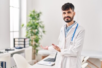 Young hispanic man wearing doctor uniform doing welcome gesture with hands at clinic