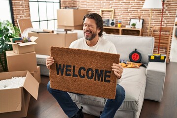 Handsome middle age man holding welcome doormat at new home winking looking at the camera with sexy...