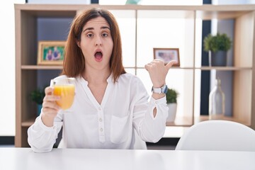 Brunette woman drinking glass of orange juice surprised pointing with hand finger to the side, open mouth amazed expression.