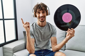 Young hispanic man holding vinyl disc wearing headphones smiling happy pointing with hand and...