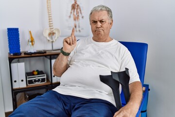Senior caucasian man at physiotherapy clinic holding crutches pointing up looking sad and upset,...