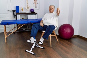 Senior caucasian man at physiotherapy clinic using pedal exerciser looking unhappy and angry showing rejection and negative with thumbs down gesture. bad expression.